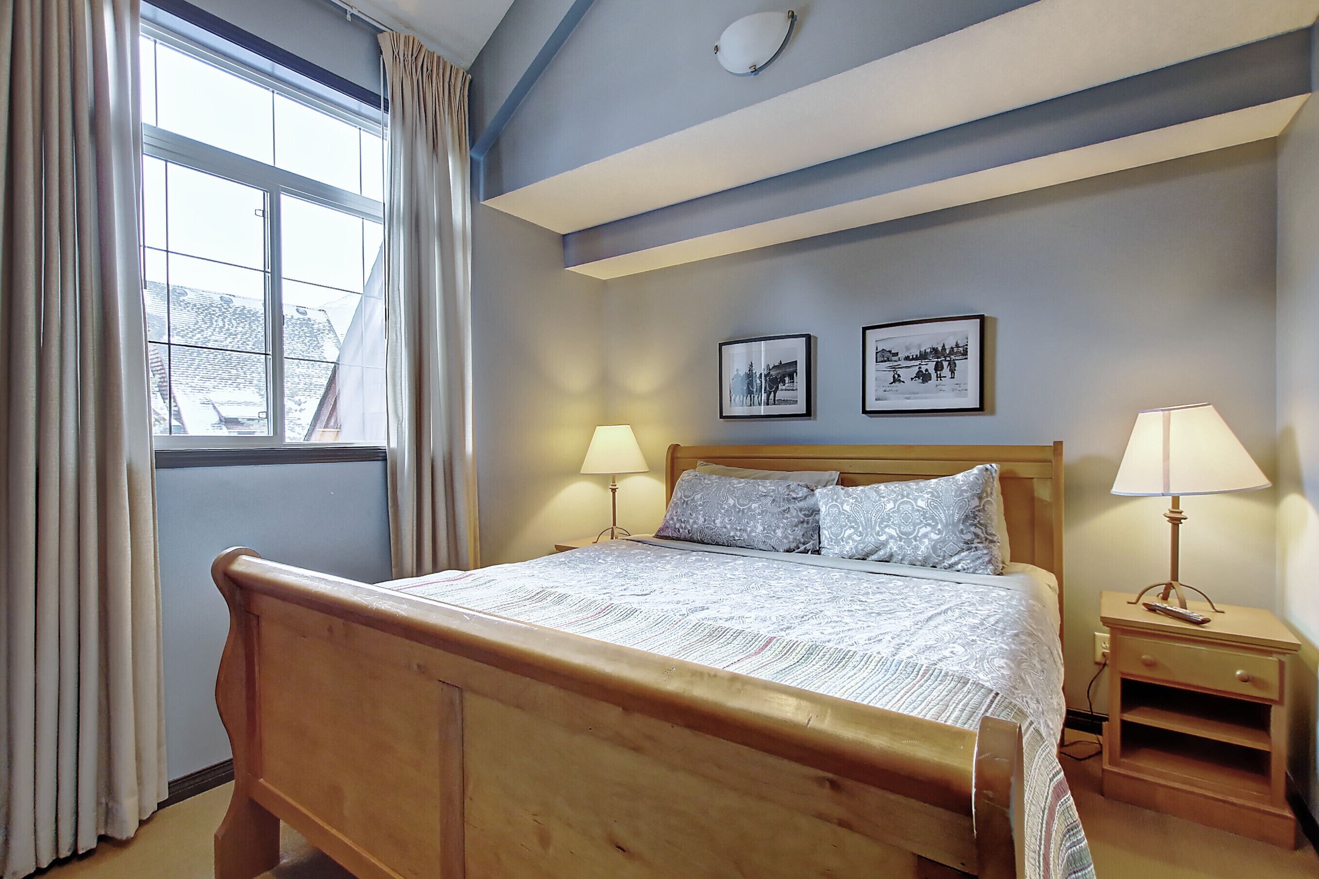A bedroom with a wooden bed, two bedside tables with lamps, a window with curtains, and two framed pictures on the wall.