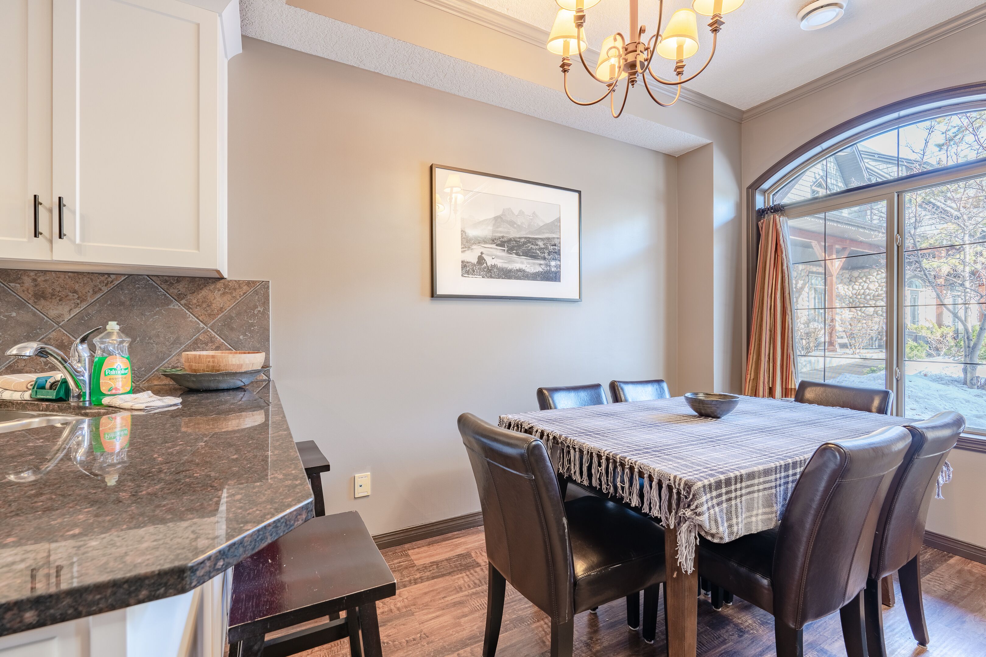 A dining area with a table set for four, a kitchen counter with a sink and dish soap, and a framed picture on the wall.