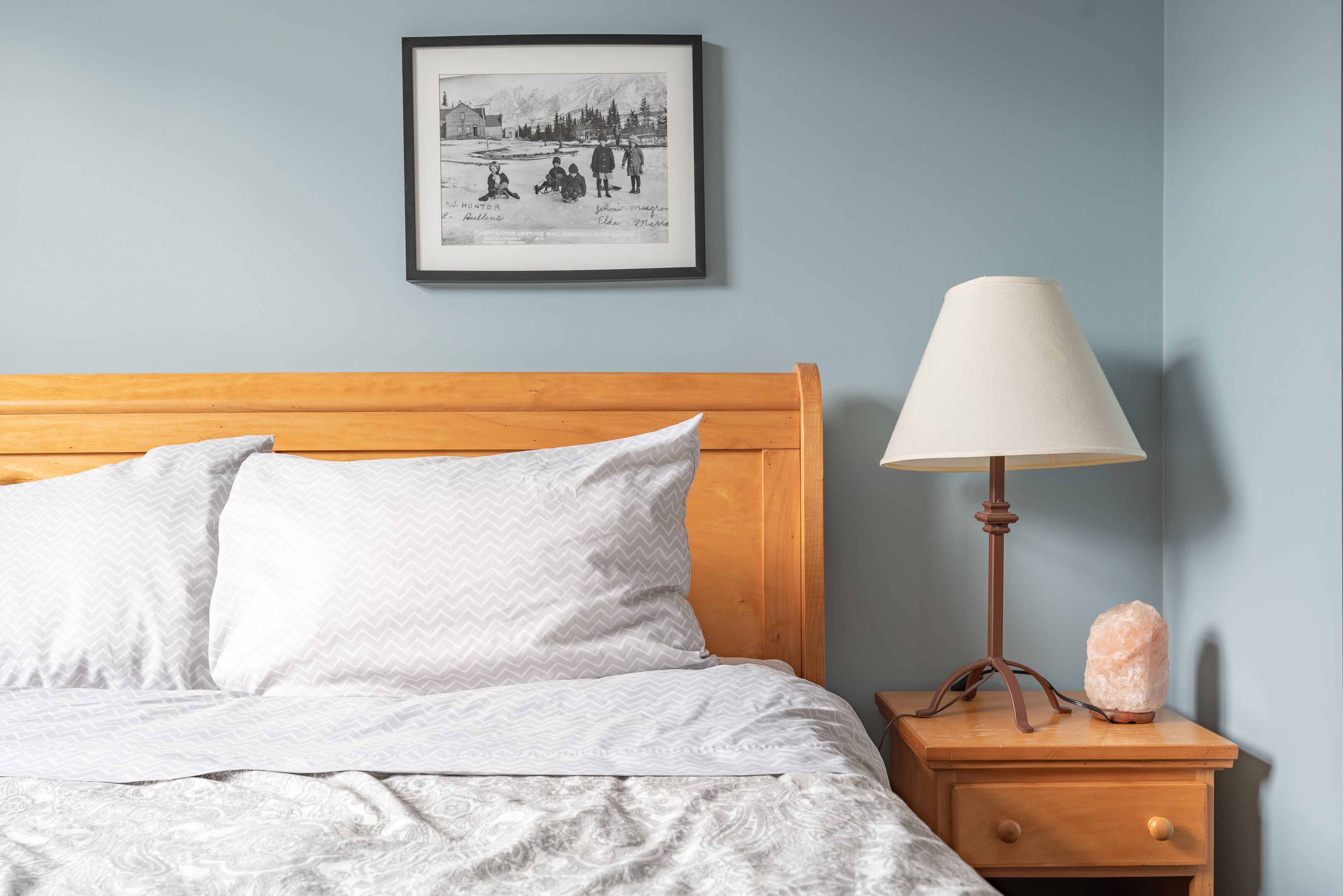 Een slaapkamer met een houten hoofdbord, beddengoed met een wit patroon, een nachtkastje met een lamp en een zoutlamp, en een ingelijste foto aan de muur.