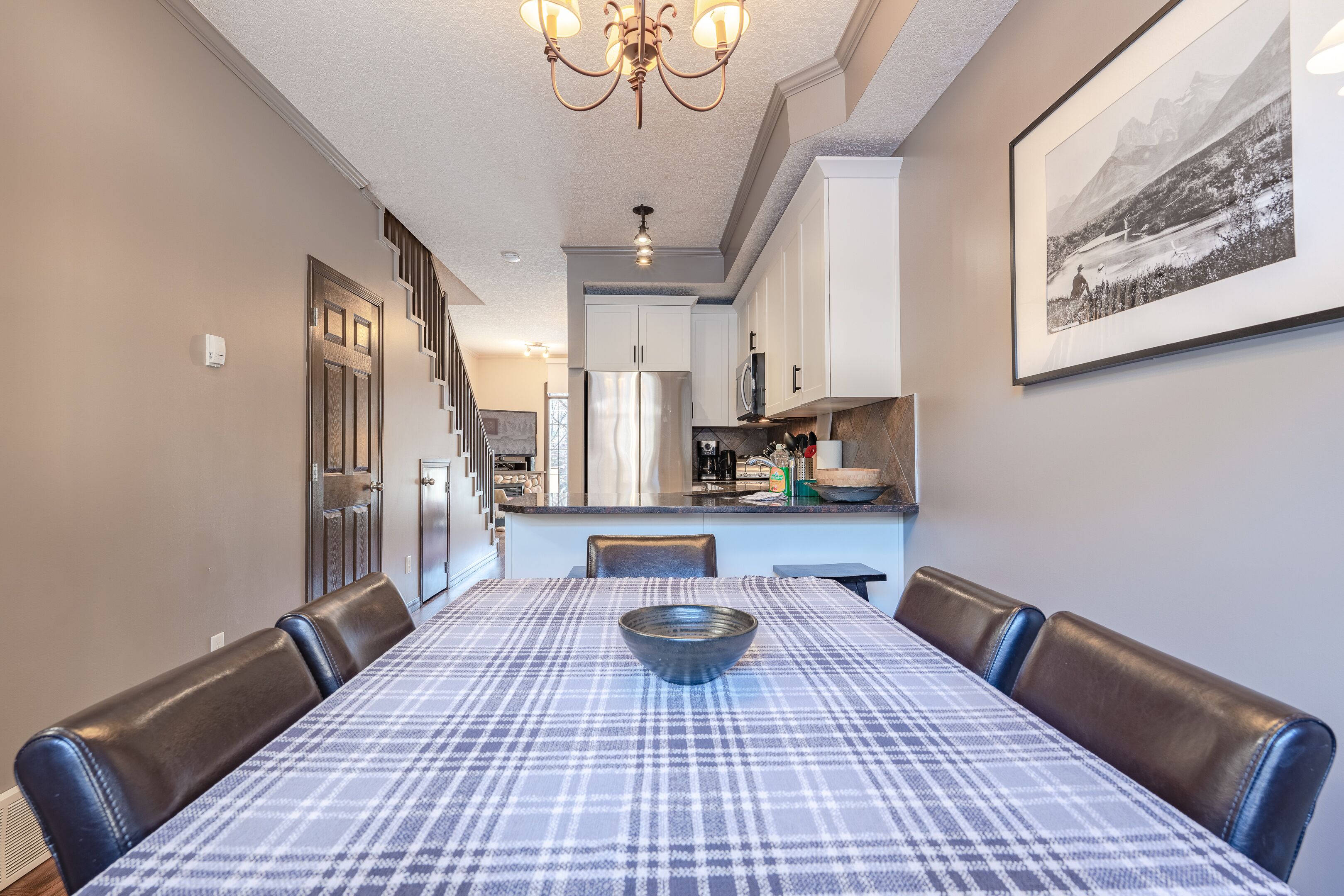 A dining area with a table set for six, a chandelier, and a kitchen in the background.