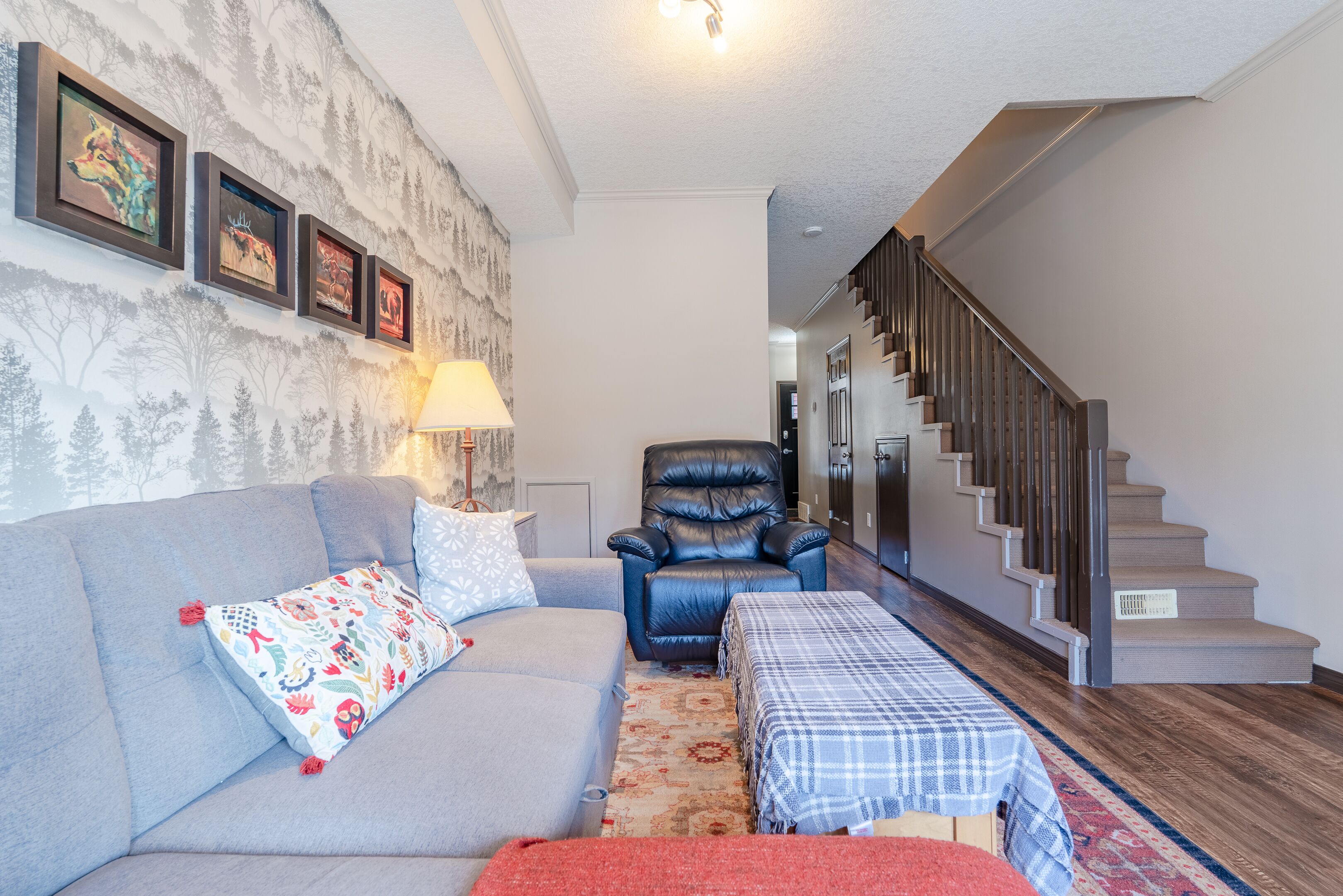 A living room with a grey sofa, a black leather chair, a patterned throw pillow, a plaid table runner, and a staircase leading to an upper level.