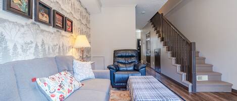A living room with a grey sofa, a black leather chair, a patterned throw pillow, a plaid table runner, and a staircase leading to an upper level.