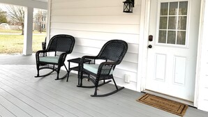 En veranda med to gyngestole og et bord, en hvid dør med et vindue, samt en sort lygte på væggen.