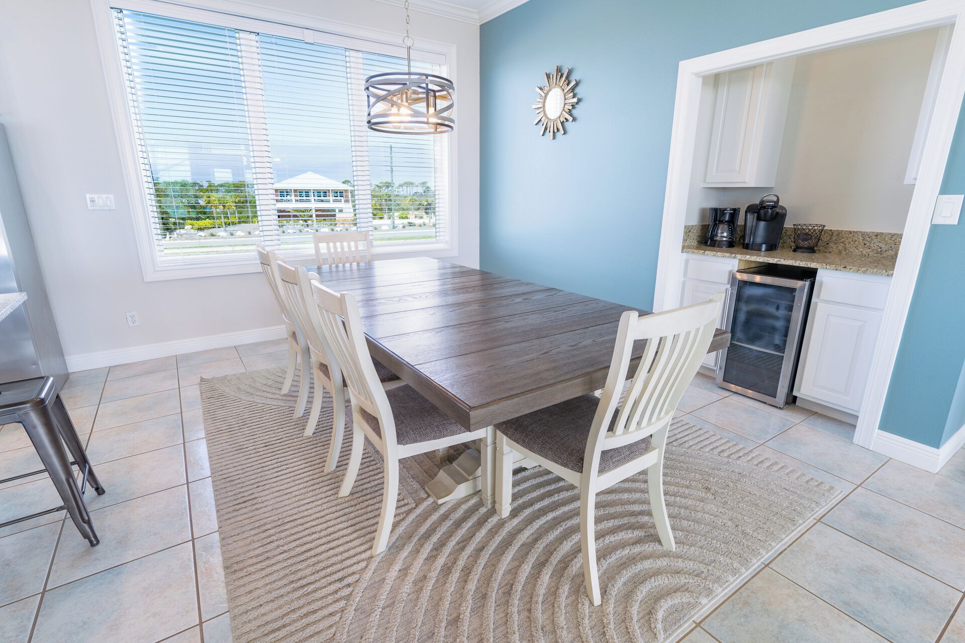 Un coin repas avec une table en bois et des chaises blanches, une cuisine avec des meubles blancs et une vue sur une maison à travers de grandes fenêtres.