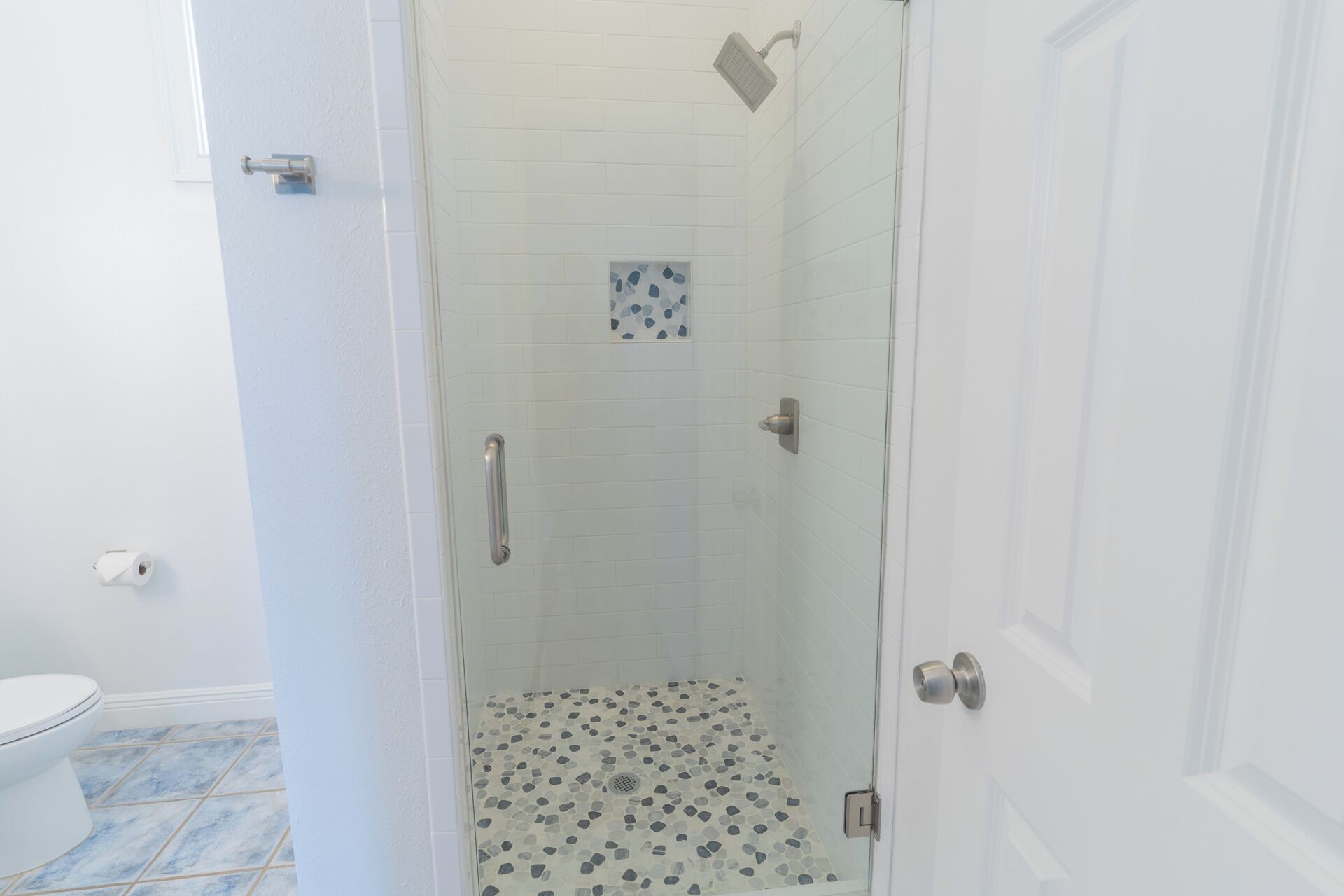 Eine gläserne Dusche mit einem Boden aus Kieselsteinen, eine weiße Tür und eine sichtbare Toilette im angrenzenden Raum.