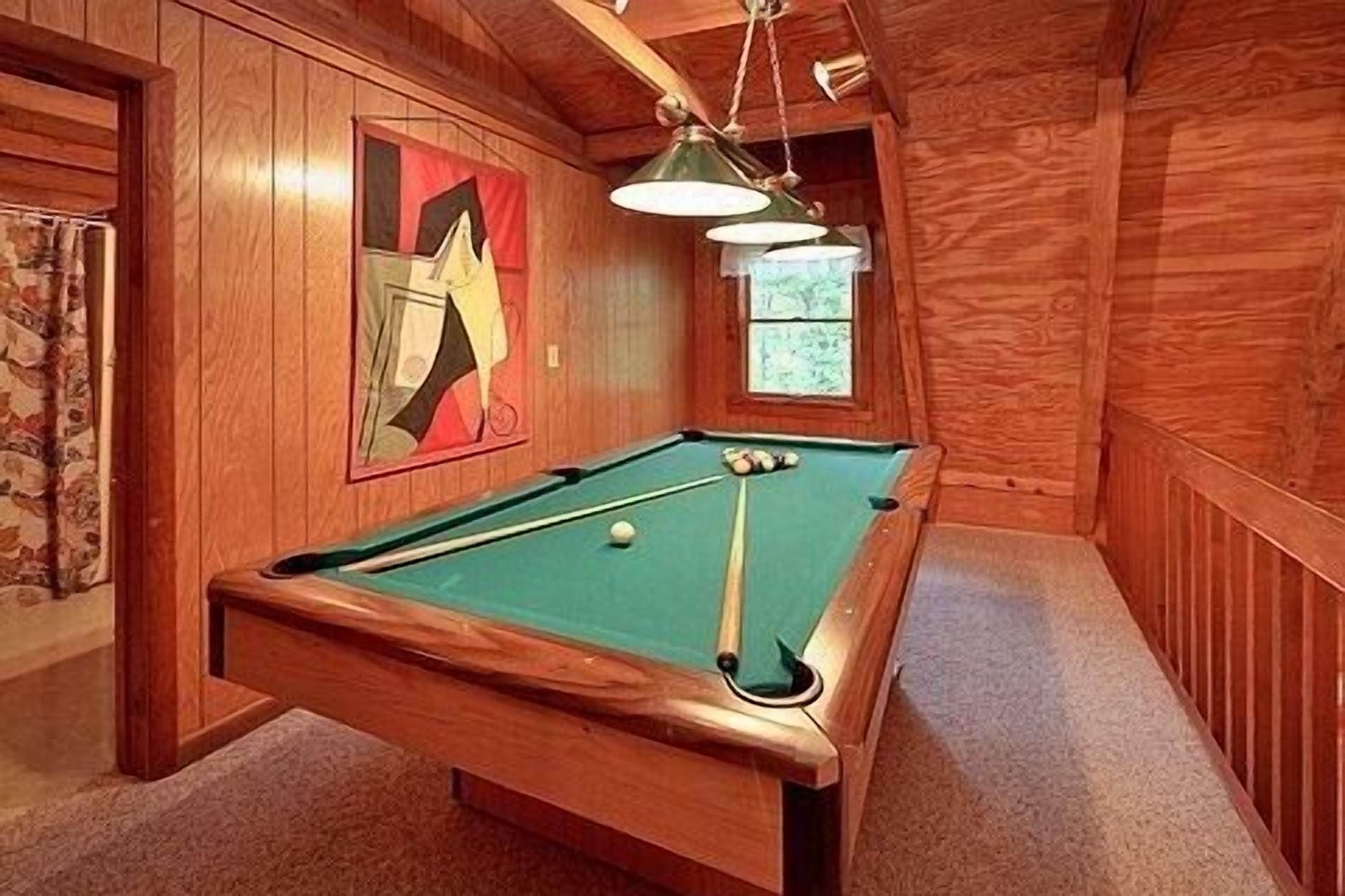 Una mesa de billar en una habitación de madera con un cuadro abstracto enmarcado en la pared y una ventana.