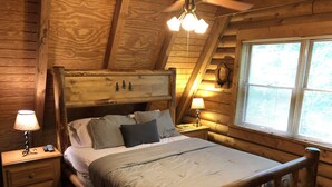 Un dormitorio de cabaña de madera con cama, mesitas de noche, lámpara y ventana.