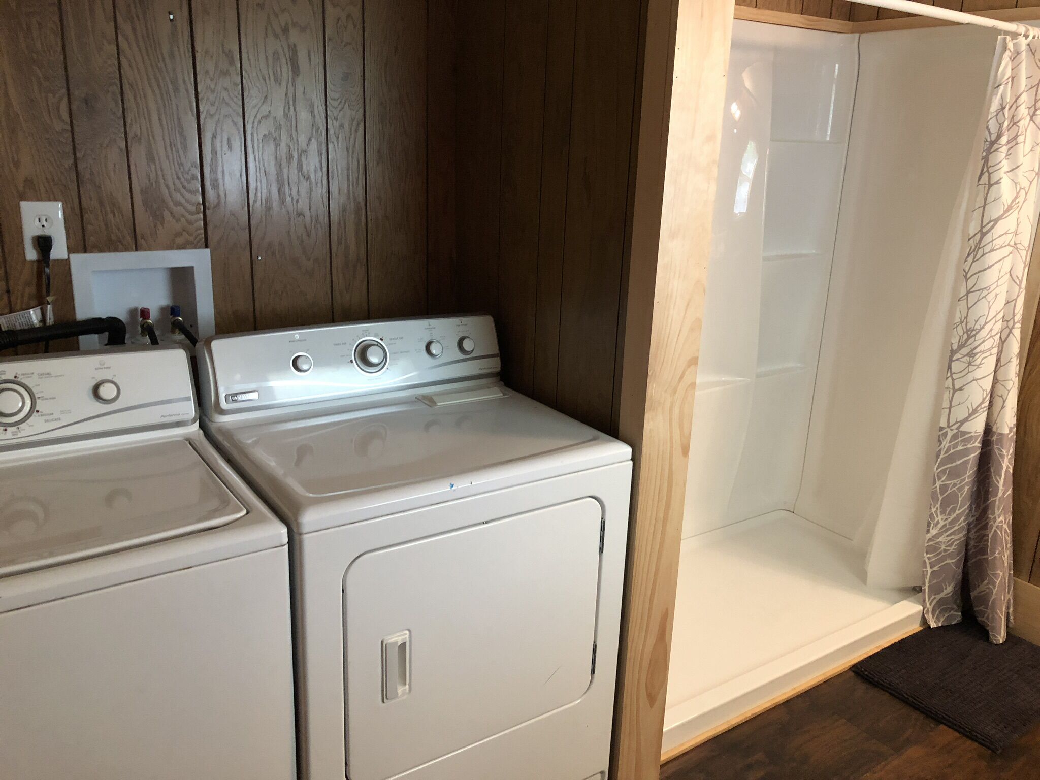 A laundry area with two white dryers, a wooden wall, and a shower with a curtain.