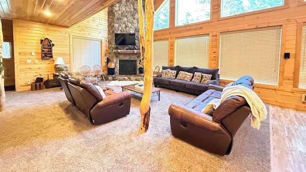 A log cabin interior with a stone fireplace, wooden walls, and large windows. - 22 MI to YNP | Firepit & Patio | Spacious Lodge (Island Park)
