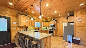 A wooden cabin interior with a kitchen featuring a granite countertop, wooden bar stools, a stainless steel refrigerator, and a built-in oven. - 22 MI to YNP | Firepit & Patio | Spacious Lodge (Island Park)