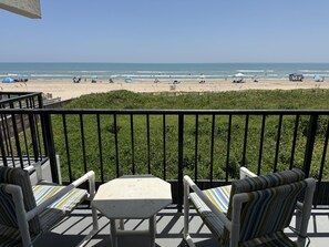 A balcony with a view of the beach, featuring a table and two striped chairs. - Oceanfront Studio, sleeps 4 -Private balcony, pool, hot tub, BBQ (South Padre Island)