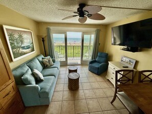 A living room with a sofa, a chair, a television, and a balcony with a view of the beach.