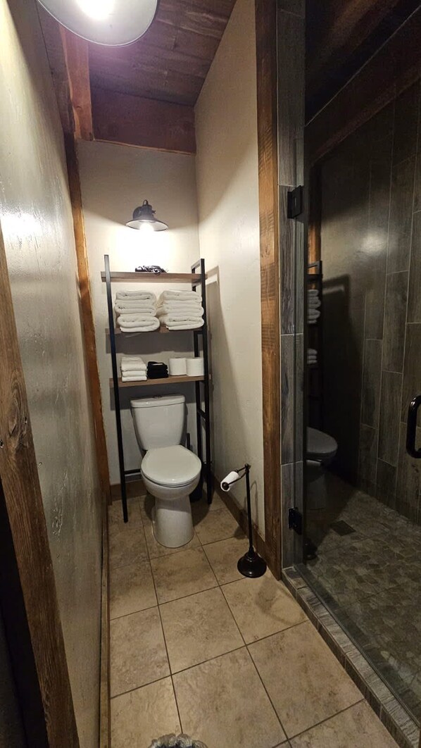 A narrow bathroom with a toilet, a shelf with towels, and a shower area. - Lookout Tower: Sleeps 4- Water Views- Near Glacier (Coram)
