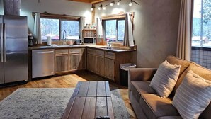 A cozy cabin interior with a wooden ceiling, a kitchen with a refrigerator and sink, a wooden dining table, and a sofa with cushions.