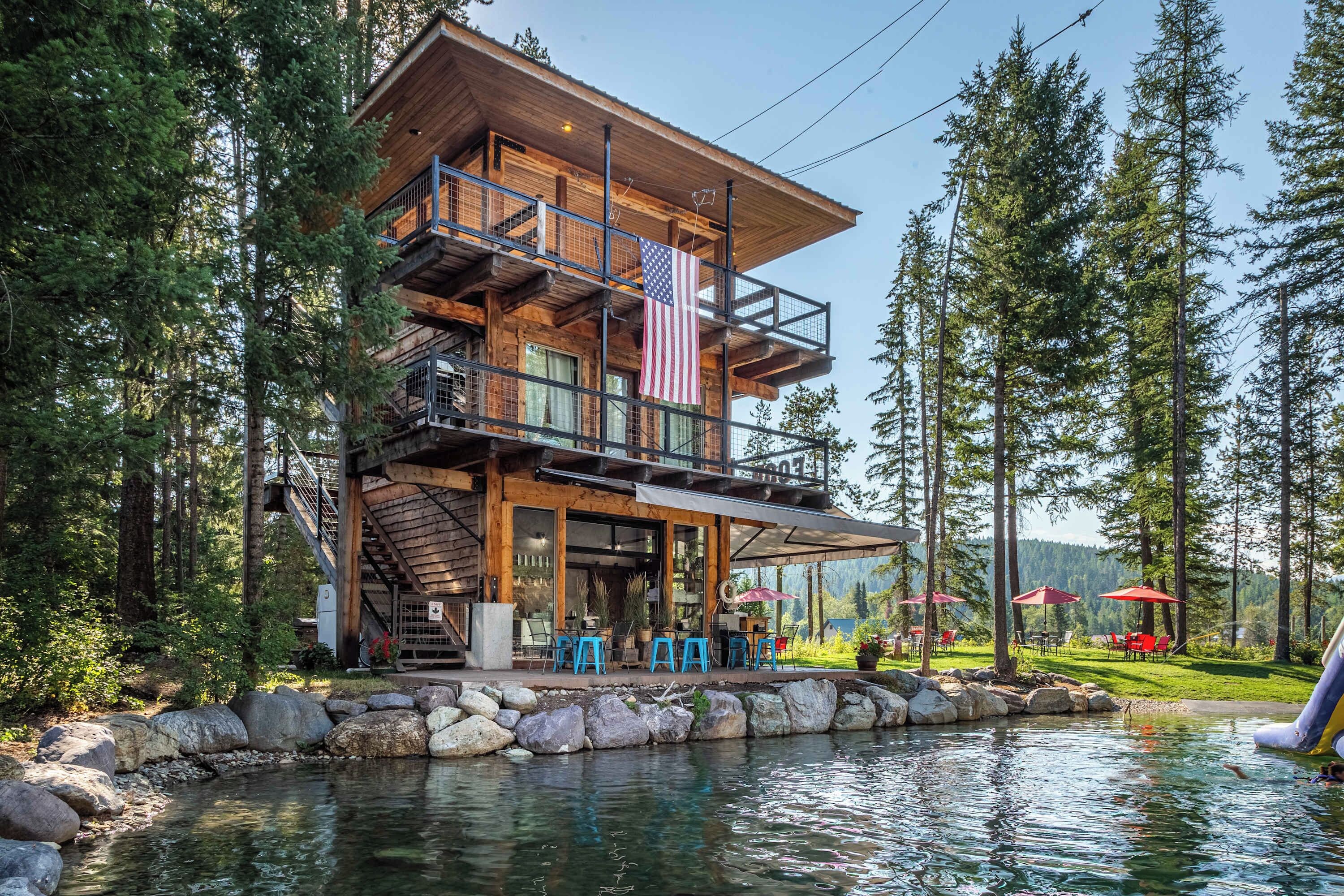 A multi-story wooden cabin with a deck, outdoor seating, and a view of a lake surrounded by trees.