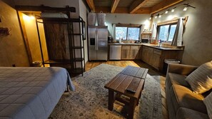 A cozy room with a bed, a wooden table, a sofa, a kitchen area with a refrigerator, and a window with curtains. - Lookout Tower: Sleeps 4- Water Views- Near Glacier (Coram)