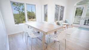 A spacious room with a large wooden table and white chairs, large windows with a view of trees, and a sliding glass door leading to an outdoor area. - Secluded 4BR/3.5BA, inside St. George Plantation (St. George Island)