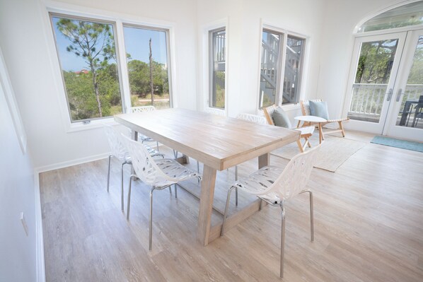 A spacious room with a large wooden table and white chairs, large windows with a view of trees, and a sliding glass door leading to an outdoor area. - Secluded 4BR/3.5BA, inside St. George Plantation (St. George Island)
