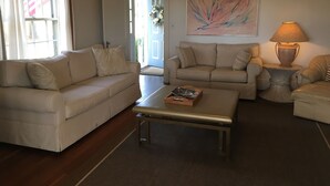 A living room with a beige sofa, a coffee table, and a lamp. There is a door leading outside and a painting on the wall.