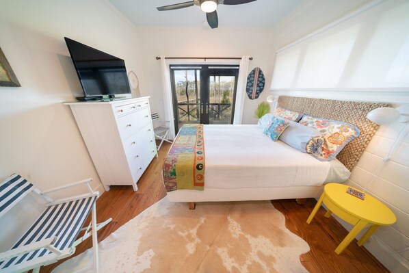 A bedroom with a bed, a TV on a dresser, a small chair, a yellow side table, and a view of the outdoors. - Bayfront, Handicap-Friendly, Pool & Elevator, Kayaks (St. George Island)