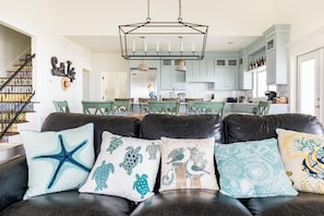 A living room with a black leather sofa, decorative pillows, a chandelier, and a view into a kitchen with light blue cabinets.