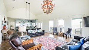 A spacious living room with a large chandelier, a patterned rug, and a modern kitchen in the background. - Bach Haus: 4  BR, 3  BA House in Gilchrist, Sleeps 18 (Bolivar Peninsula)