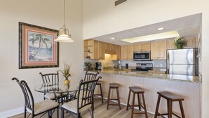 A kitchen with wooden cabinets, a stainless steel refrigerator, a microwave, and a coffee maker. There is a dining table with chairs and a bar stool area.
