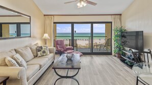 A living room with a sofa, a coffee table, a TV, and a balcony with a view of the ocean.