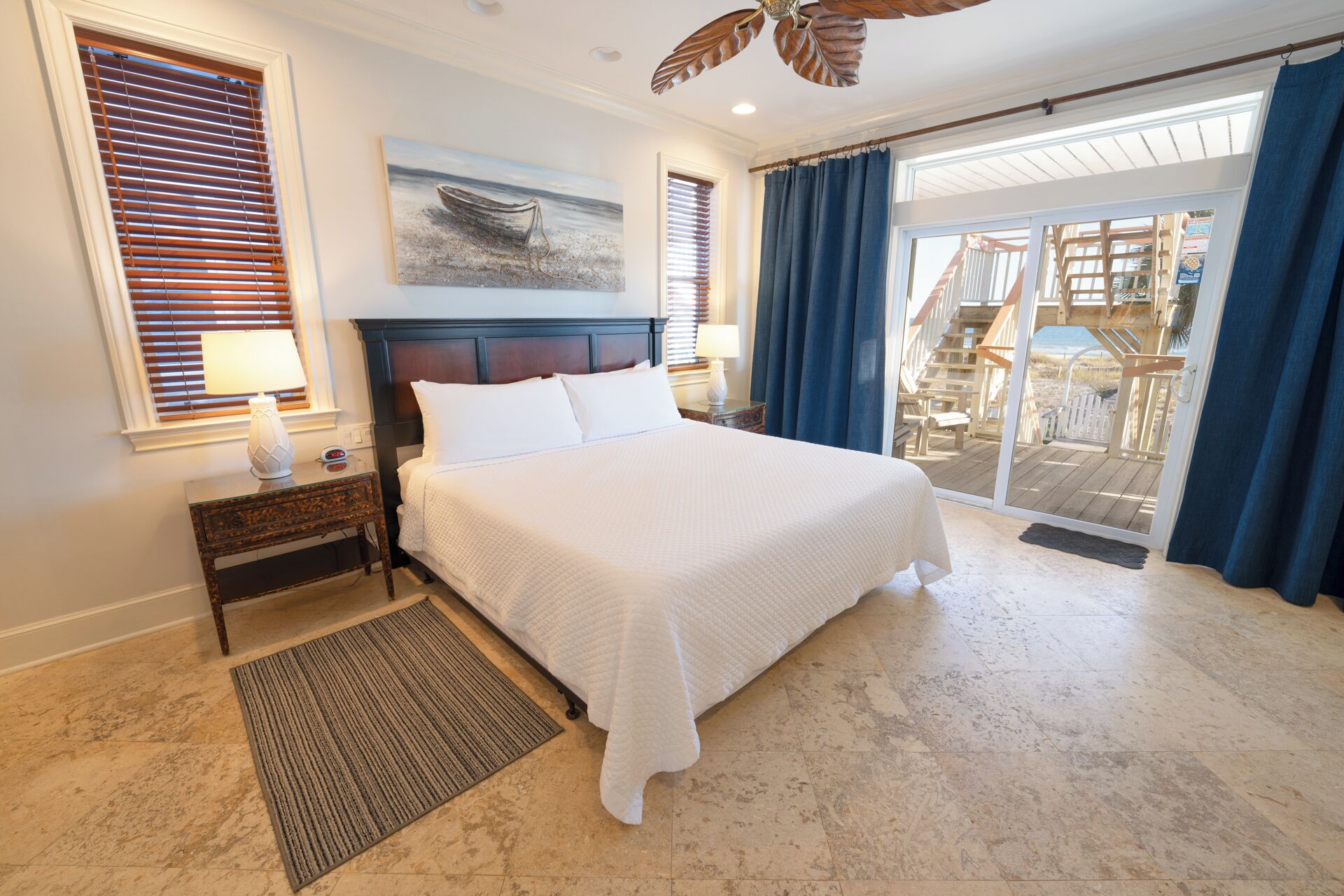 Une chambre avec un grand lit, une tête de lit en bois, un tableau représentant un bateau, une table de chevet avec une lampe et une vue sur une terrasse en bois, visible à travers une porte vitrée.