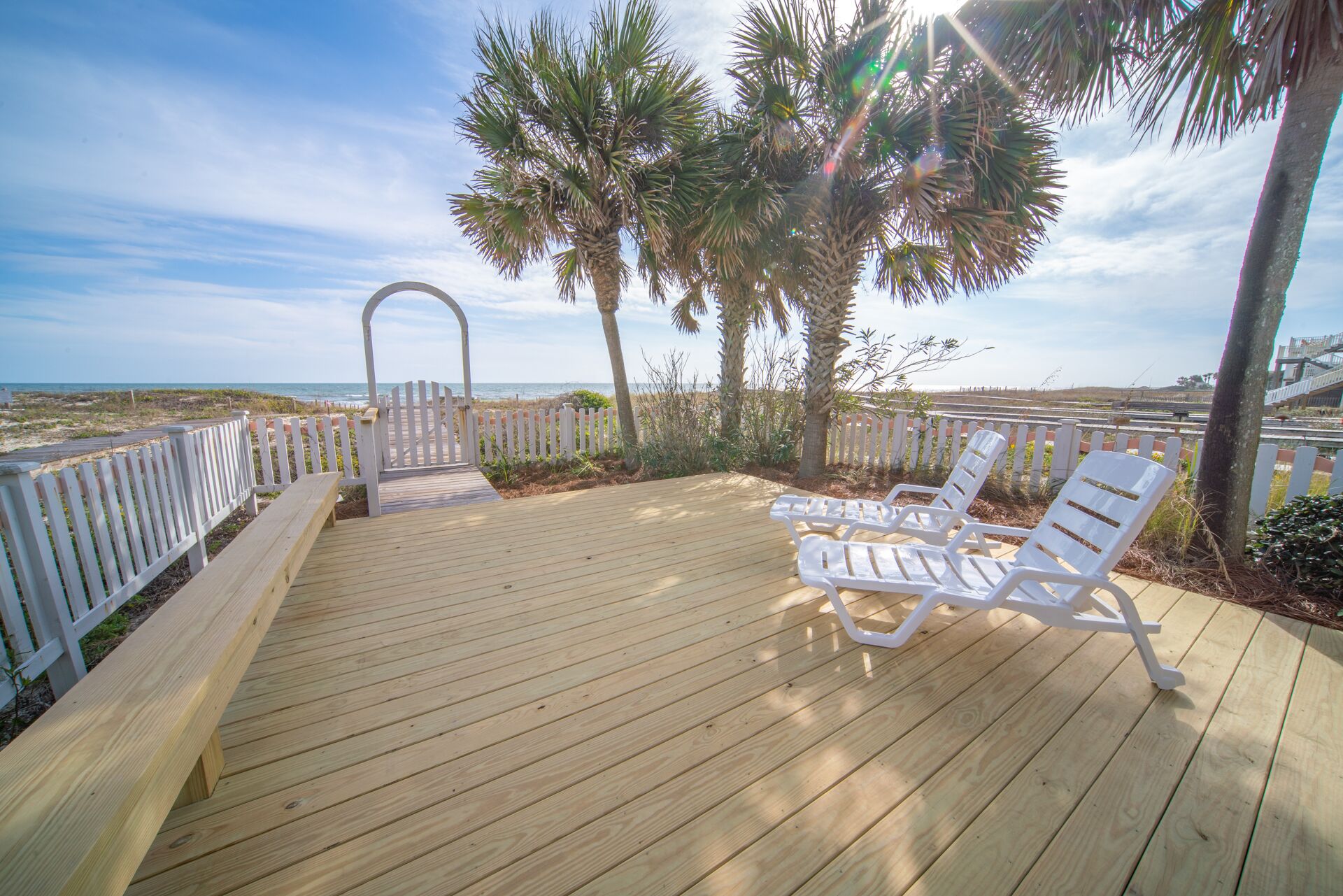 A wooden deck with white railings, two white lounge chairs, and a view of the ocean.