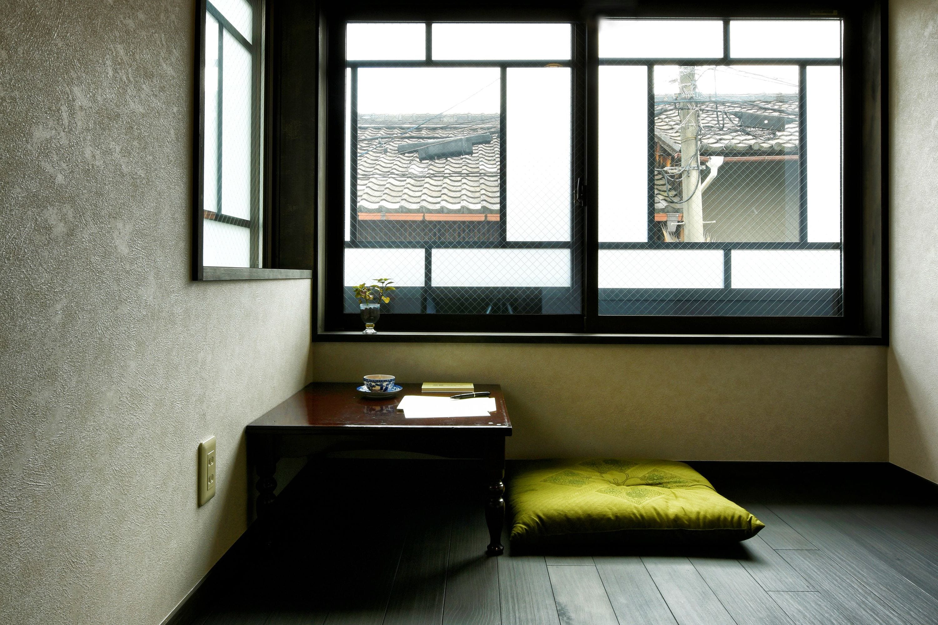 A corner room with a large window, a small table with a bowl and a cup, and a green cushion on the floor.