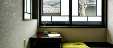 A corner room with a large window, a small table with a bowl and a cup, and a green cushion on the floor.
