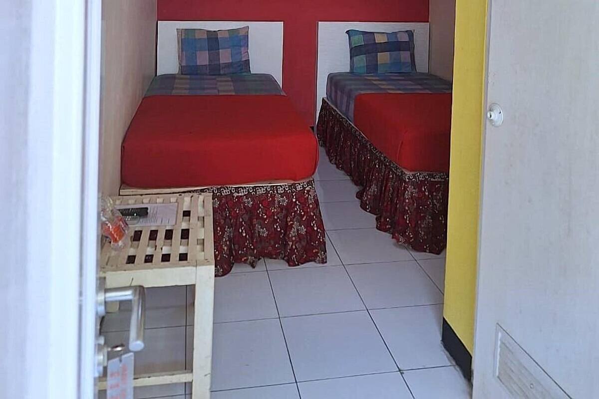 A small, narrow room with two beds, each with a red bedspread and matching pillows. The walls are painted red, and there is a small framed picture on the wall. The floor is tiled, and there is a door on the right side.