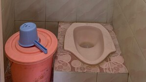 A small, tiled bathroom with a toilet, a blue bucket, and a pink container. - Ria Hotel Jember (Jember)