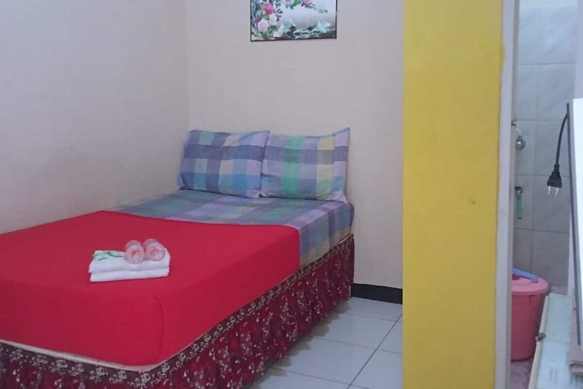 A small bedroom with a bed, a red bedspread, a floral-patterned skirt, a wall-mounted picture, and a red mat with a bear design.