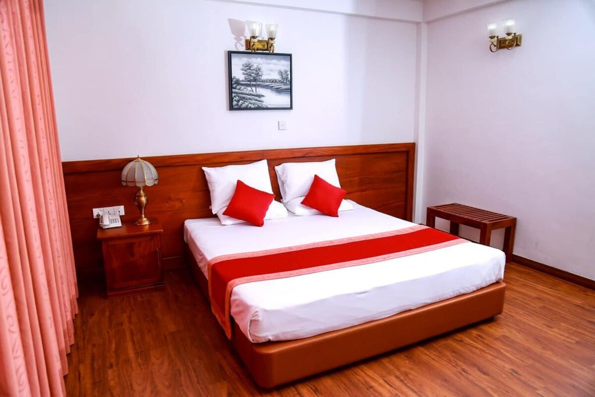 Une chambre d’hôtel avec un lit en bois, du linge de lit blanc, un plaid rouge, une table de chevet avec une lampe et un tableau au mur.