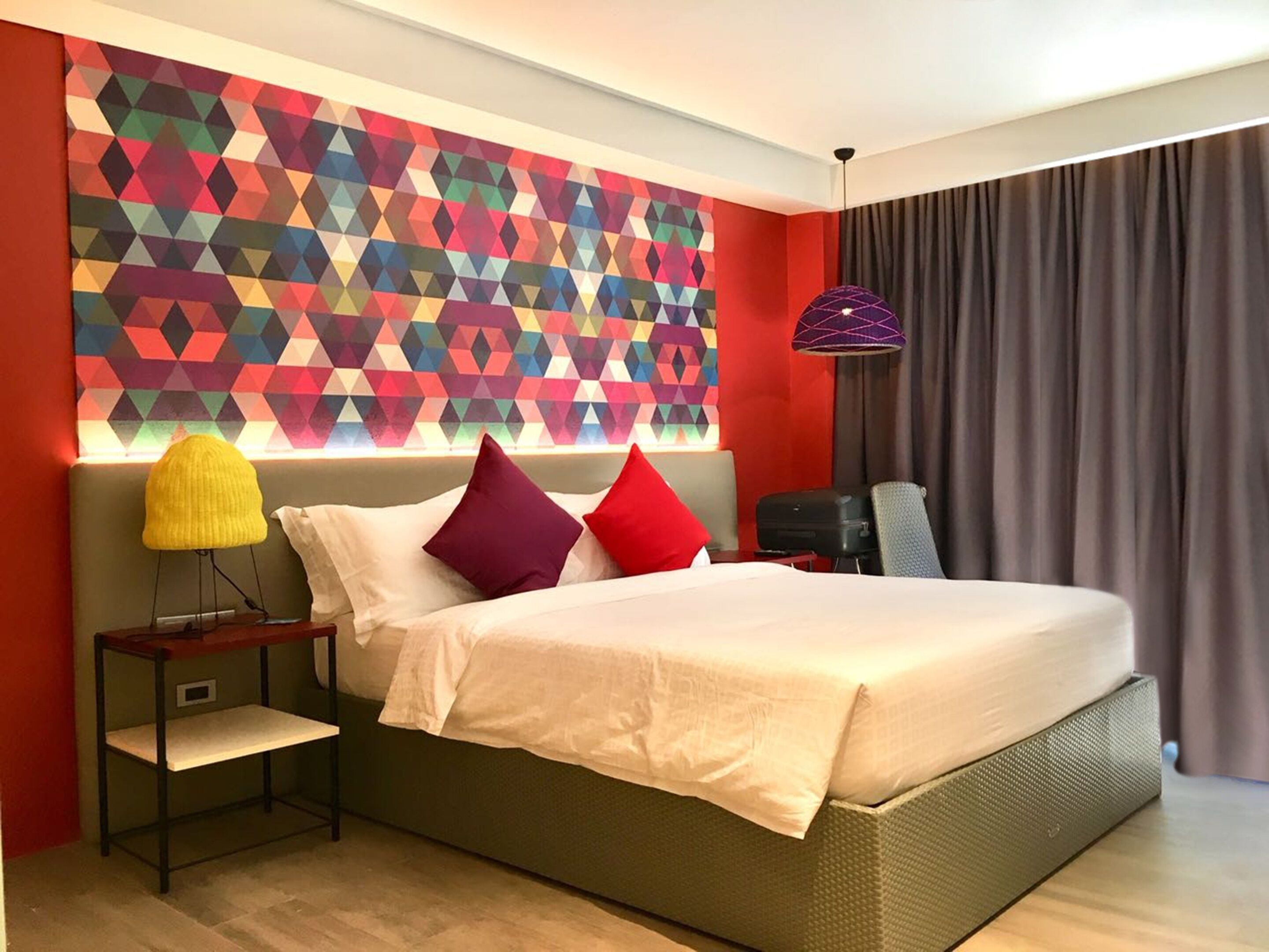 A modern hotel room with a large bed, a geometric-patterned wall, a side table with a lamp, and a view of the outside through a window with curtains.