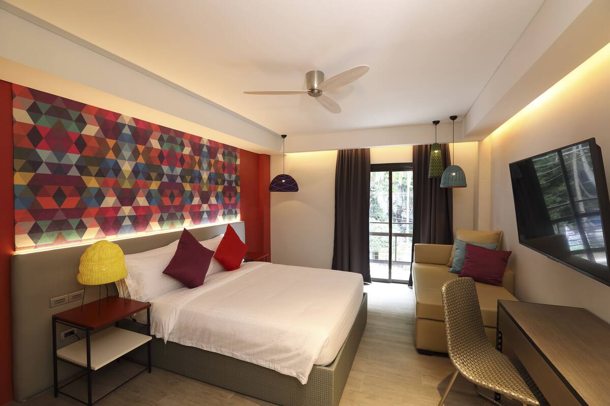 A modern hotel room with a bed, a desk, a chair, a ceiling fan, and a large window.