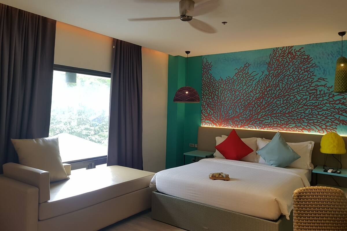 A modern hotel room with a bed, a sofa, a ceiling fan, and a wall with a coral reef mural.