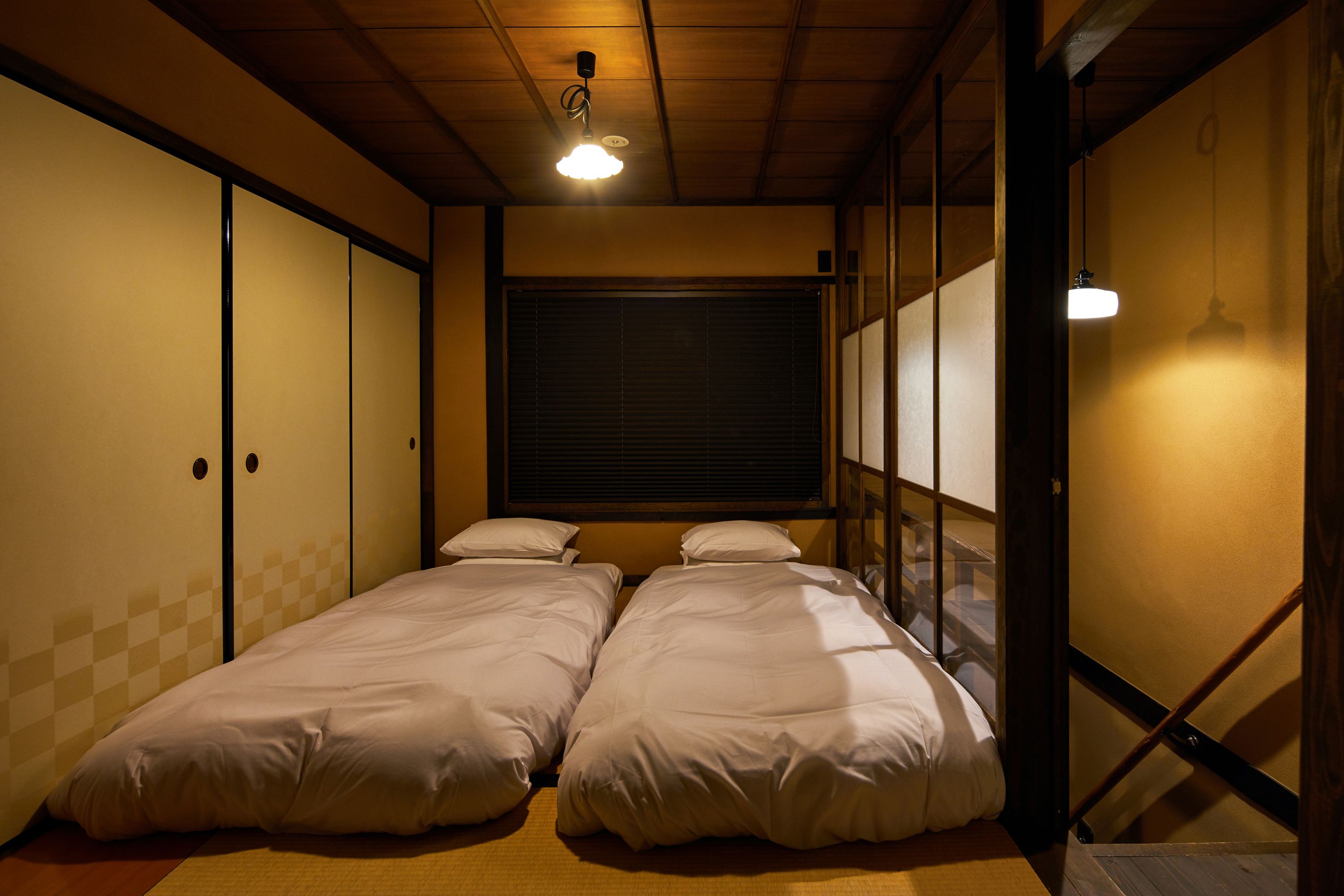 A room with two beds, a window with blinds, and a ceiling light.