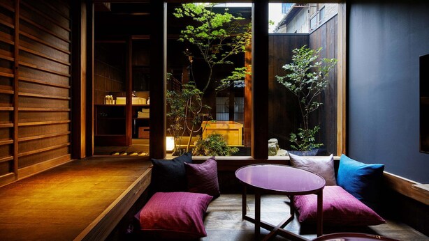 A cozy interior with a wooden bench, a round table, and cushions. A large window reveals a garden outside.