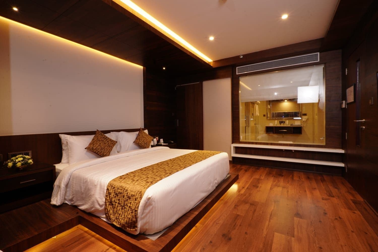 Habitación de hotel con una cama grande, suelo de madera, un banco empotrado y vista al interior del hotel.