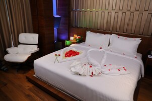 A neatly made bed with white linens and rose petals, a white armchair, and a wooden headboard.