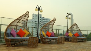 Dos sillones de mimbre con cojines, una mesita y una farola en una terraza de madera con un paisaje urbano al fondo.