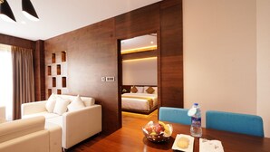 Habitación de hotel con pared de madera decorativa, sofá blanco, mesa de comedor con frutero, botella de agua y plato de galletas.