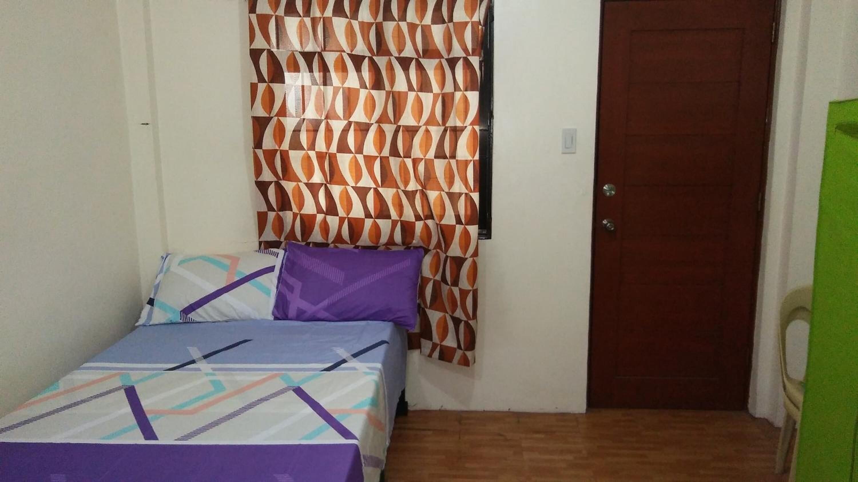 A small, single-bed room with a patterned curtain, a wooden door, and a green chair.