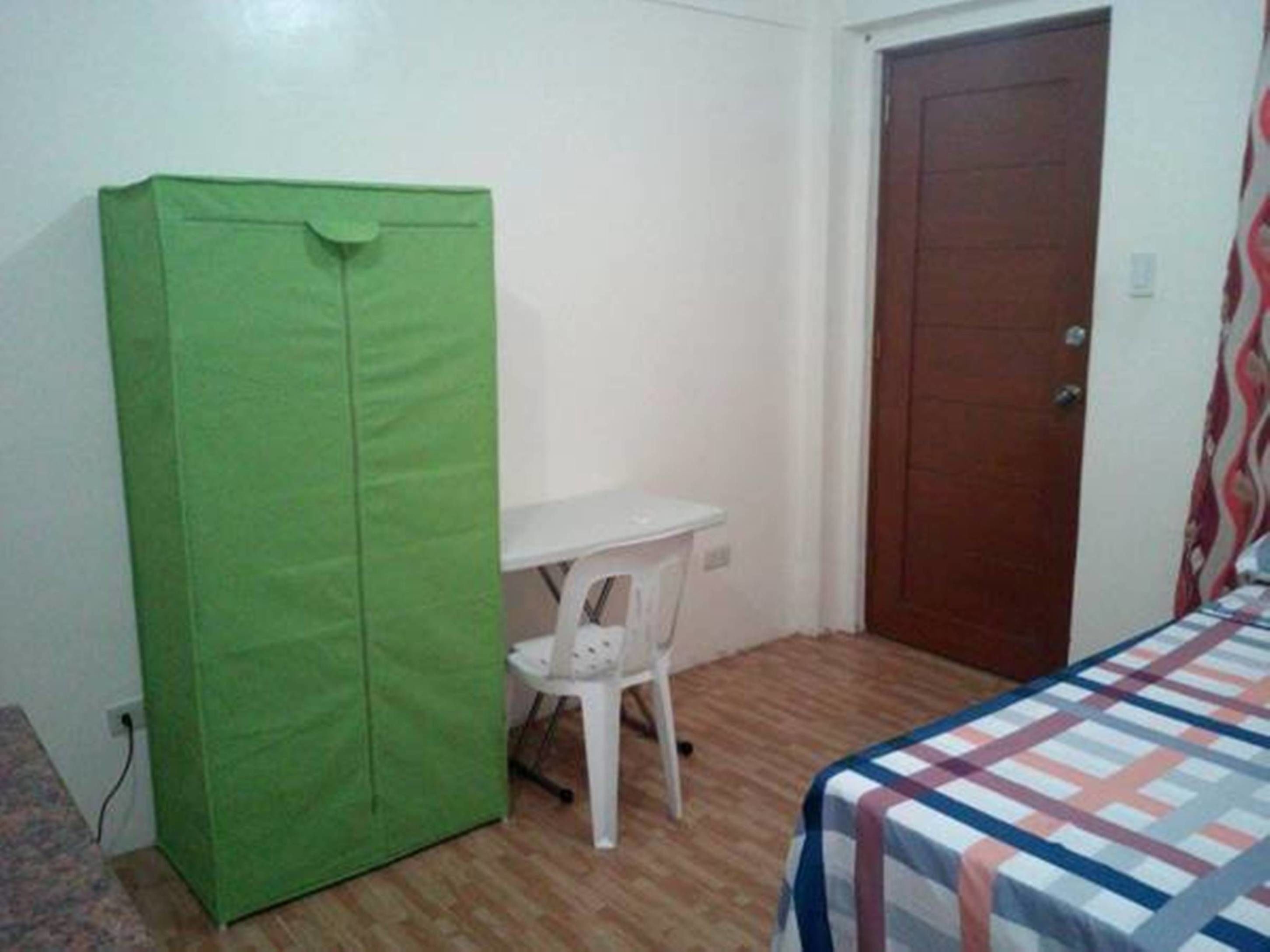 A small room with a green cabinet, a white table with a chair, a wooden door, and a bed with a patterned bedspread.