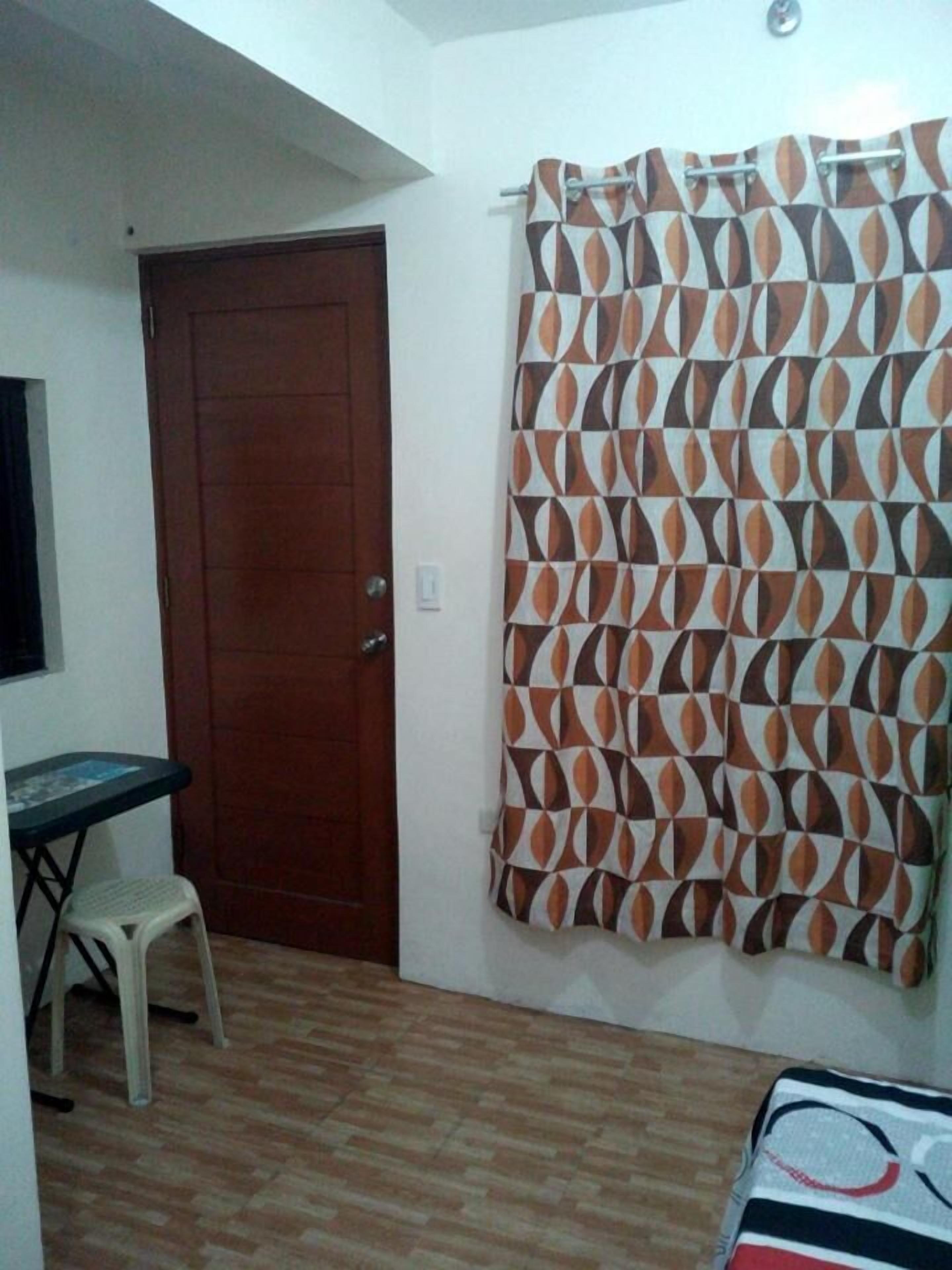 Una habitación con puerta de madera, cortina estampada, una mesita con un taburete y una cama con manta a rayas.