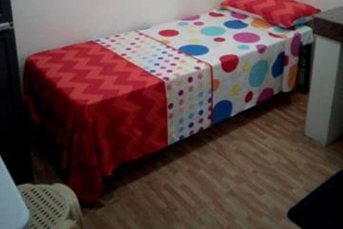 A small, single-bed room with a colorful polka dot bedspread, a red blanket, and a small desk with a chair.