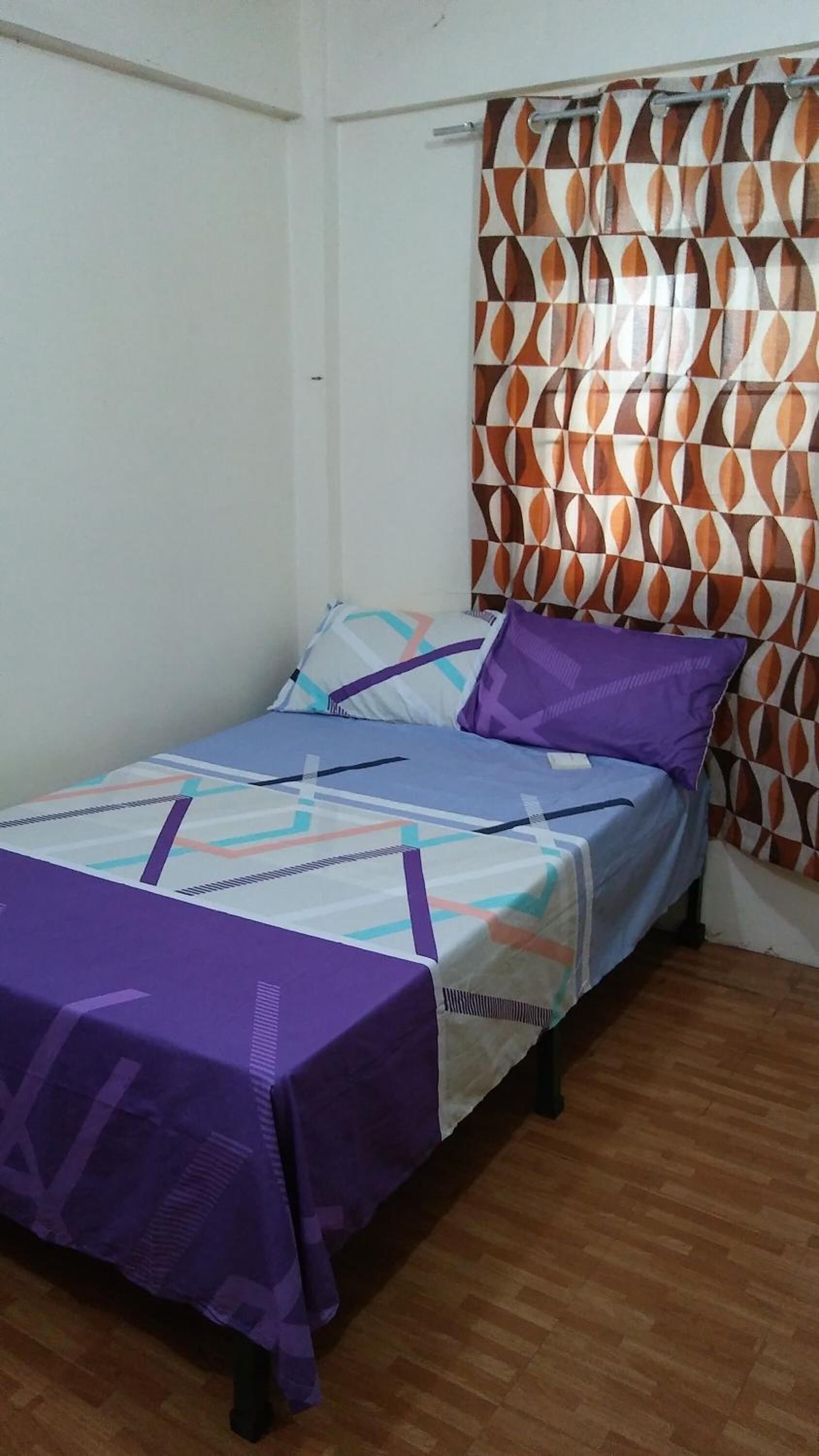 A single bed with a patterned bedspread, a purple pillow, and a curtain with a geometric design.